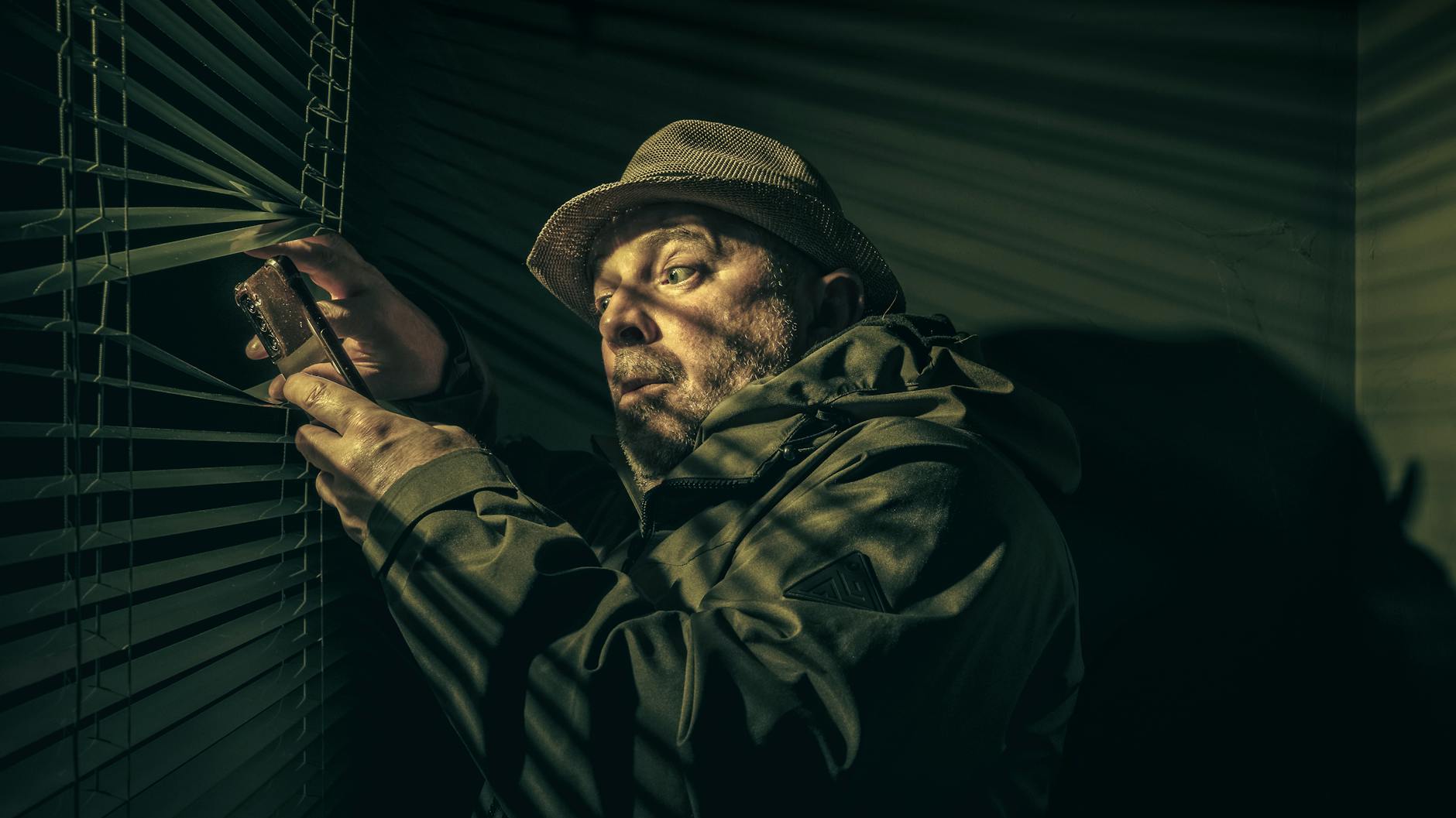 Kryminały o morderstwach wśród sąsiadów: osiedlowe tajemnice zza ściany i z klatki schodowej Mężczyzna w kapeluszu podejrzliwie zagląda przez żaluzje z telefonem w dłoni